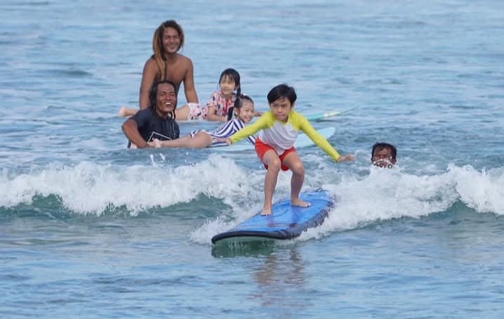 A young student in a yellow rash guard riding whitewater on a blue board, with the instructor and more kids cheering from a second board behind him