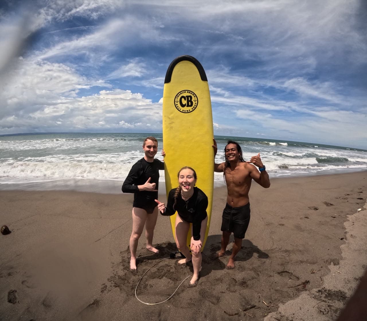 Two students and their instructor posing beachside with a yellow surfboard, throwing shakas under a bright sky