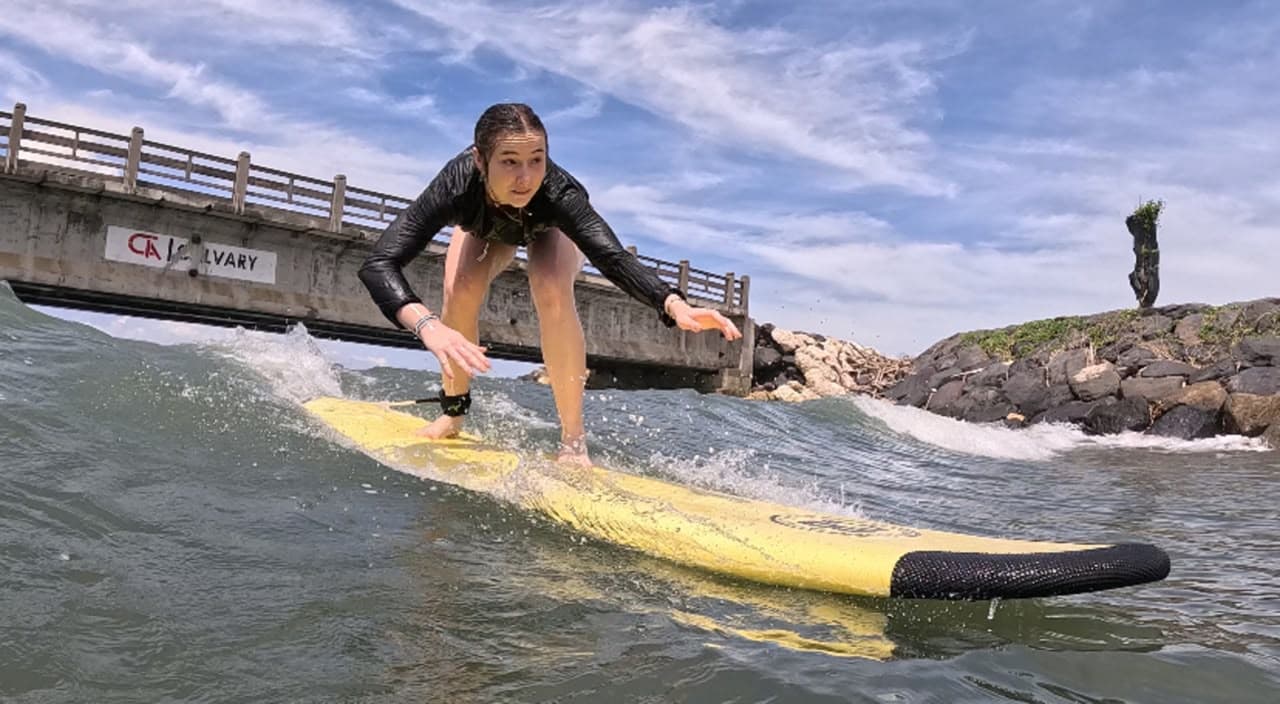 A student crouched low on a yellow board riding a small wave beside a rocky jetty and footbridge
