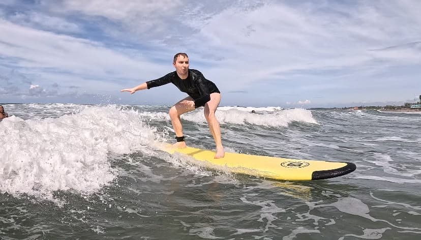 A student riding a breaking wave standing tall on a yellow board, arm out for balance, another surfer waiting behind him